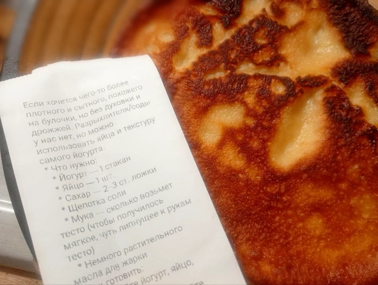 A browned yogurt pancake beside the printed Russian-language instructions for the suggested yogurt dough, photographed by the author.